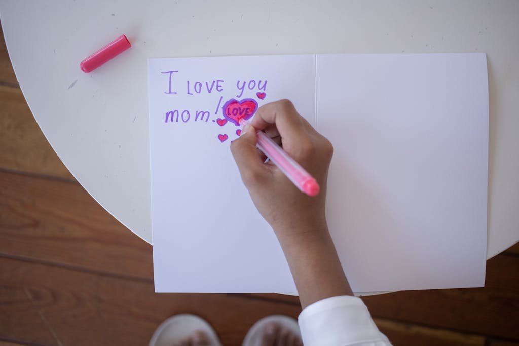 A child writes 'I love you mom' on a card with pink and purple pens on a white table.